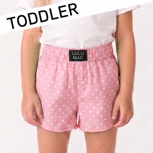 Child wearing pink polka dot shorts with 'Lulu Mac' label, white top, on light gray background.