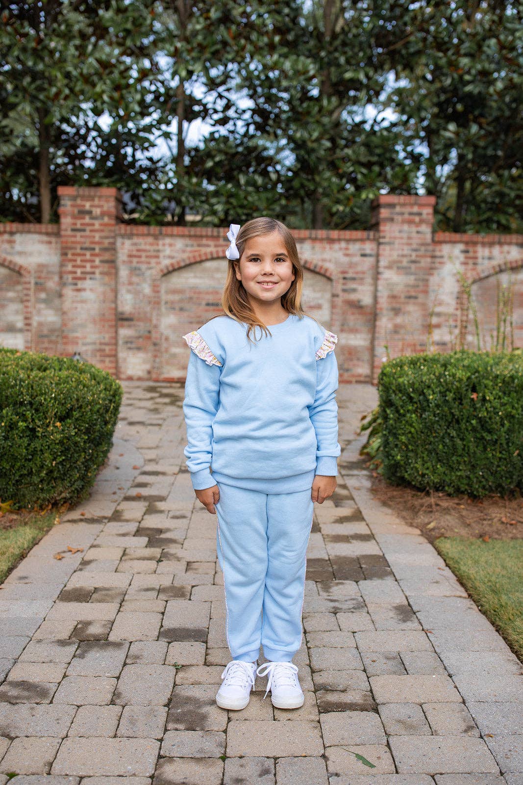 A young girl stands on a paved walkway outdoors wearing a light blue matching lounge set with white lace-trimmed shoulder details, white sneakers, and a small white bow in her hair. A brick wall and green hedges are behind her.