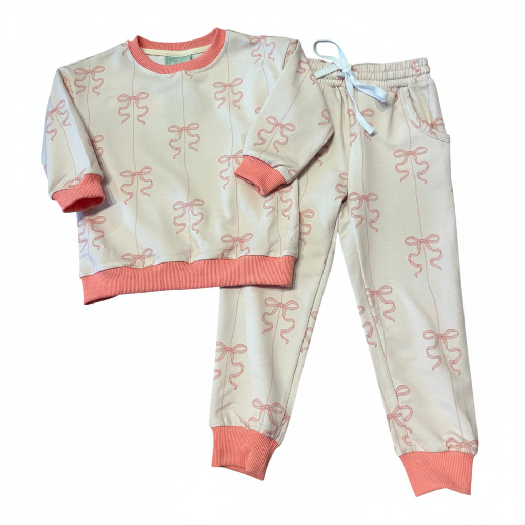 A two-piece children's activewear set featuring a long-sleeve top and matching pants. The pajamas are light beige with an all-over pink bow print, coral pink ribbed trim on the collar, cuffs, and hem, and a light blue drawstring on the pants.