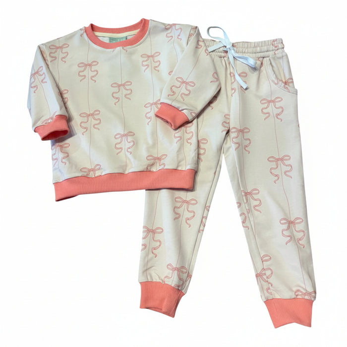 A two-piece children's activewear set featuring a long-sleeve top and matching pants. The pajamas are light beige with an all-over pink bow print, coral pink ribbed trim on the collar, cuffs, and hem, and a light blue drawstring on the pants.