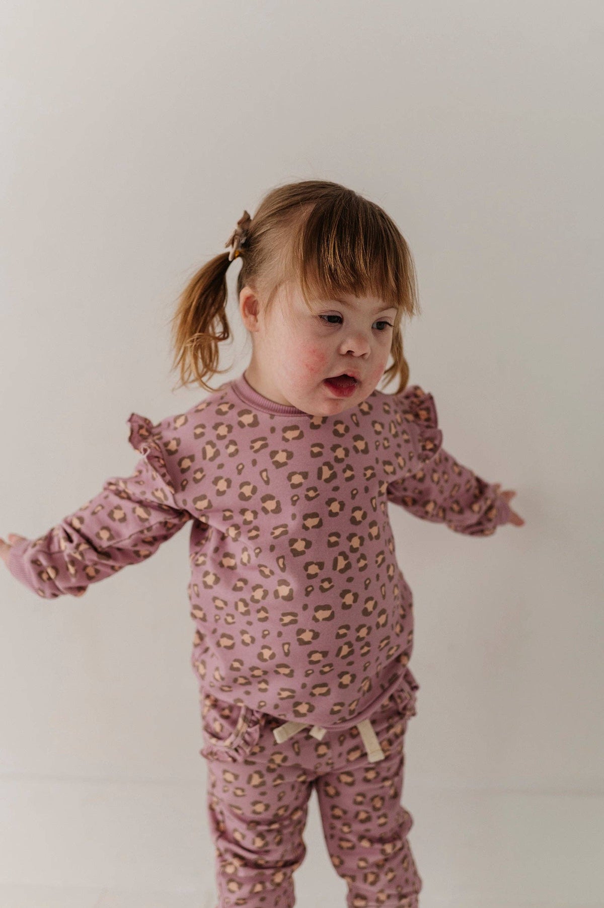A toddler girl wearing a dusty pink long-sleeve pajama set with a small beige and brown animal print, standing against a plain off-white background.
