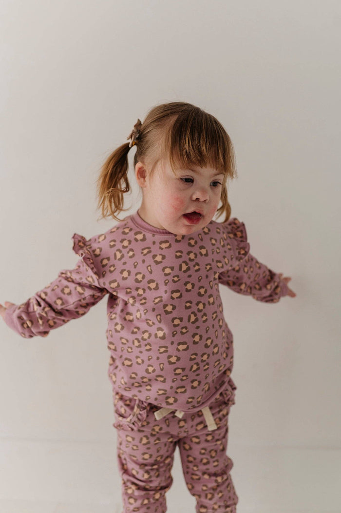 A toddler girl wearing a dusty pink long-sleeve pajama set with a small beige and brown animal print, standing against a plain off-white background.