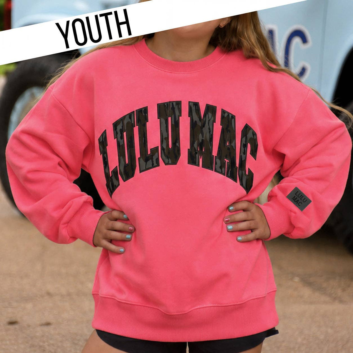 A youth model wearing a bright pink crewneck sweatshirt with 'LULUMAC' printed in black across the chest, set against a blurred outdoor background with a vehicle and a 'YOUTH' banner overlay.