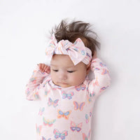 A baby lying on a white background wearing a light pink long-sleeve bodysuit with a colorful butterfly print and a matching knotted bow headband.