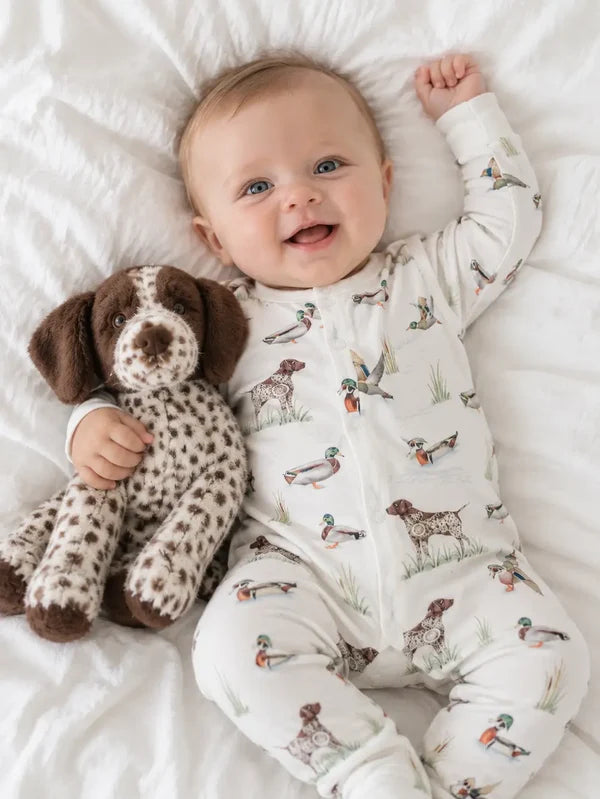 A baby lying on white bedding, wearing a white long-sleeve footless onesie printed with ducks, geese, and dogs, while holding a brown and white spotted plush dog toy.