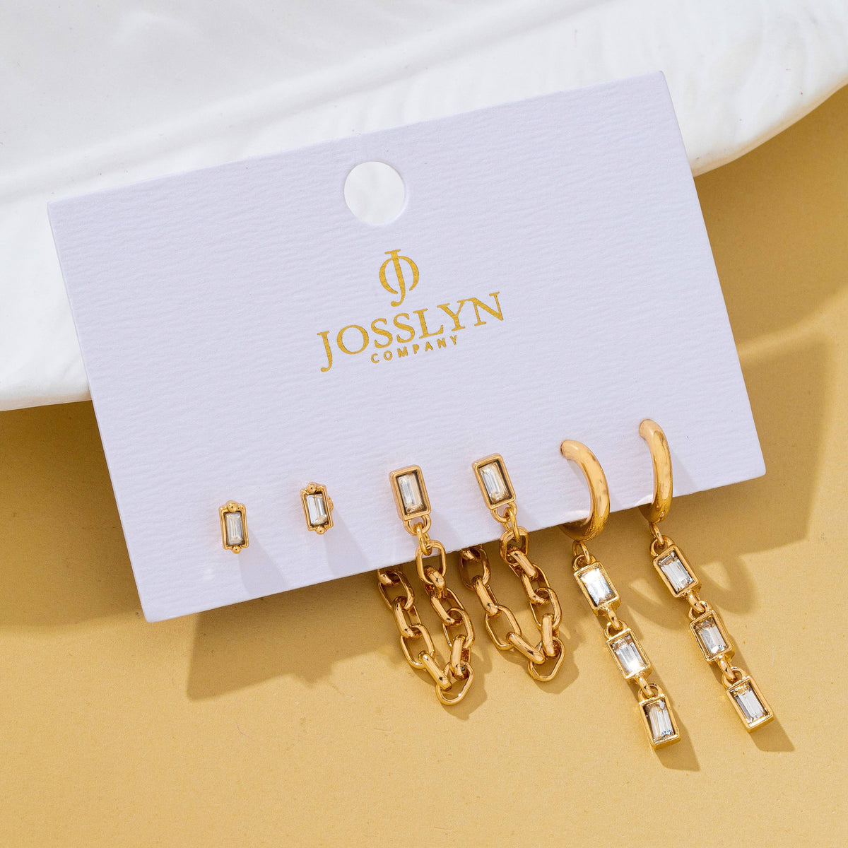 A set of six gold-tone earrings displayed on a white Josslyn Company backing card, photographed on a warm beige surface with a white fabric backdrop.