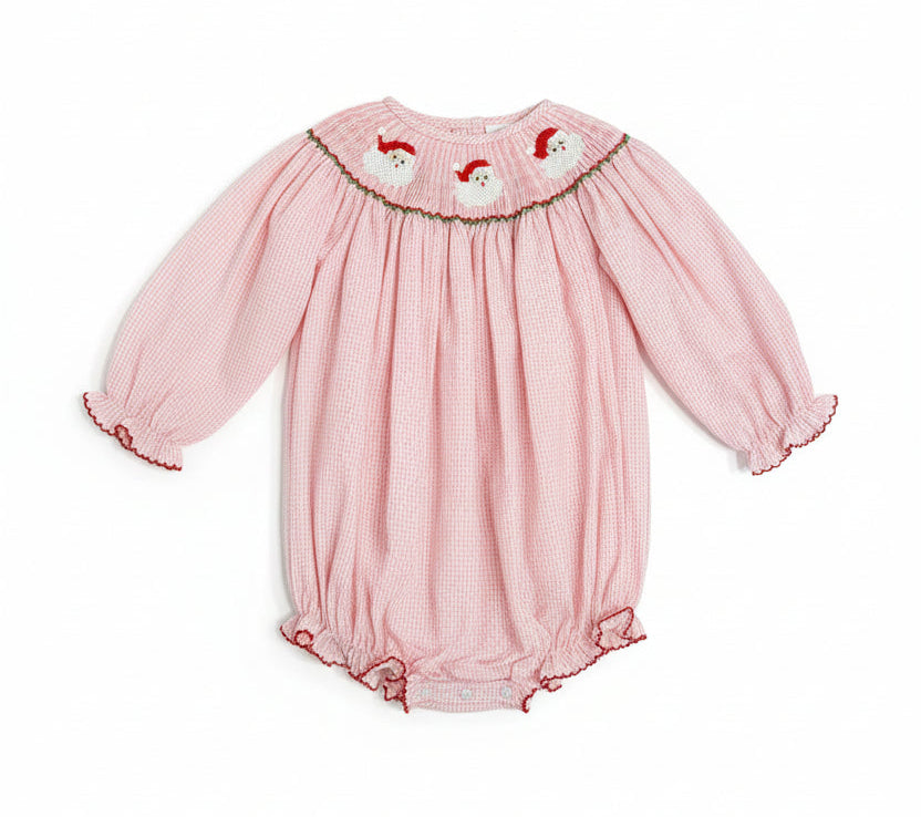 A pink and white gingham baby smocked romper with long sleeves, elastic cuffs, and a smocked Peter Pan collar decorated with three embroidered Santa Claus faces.