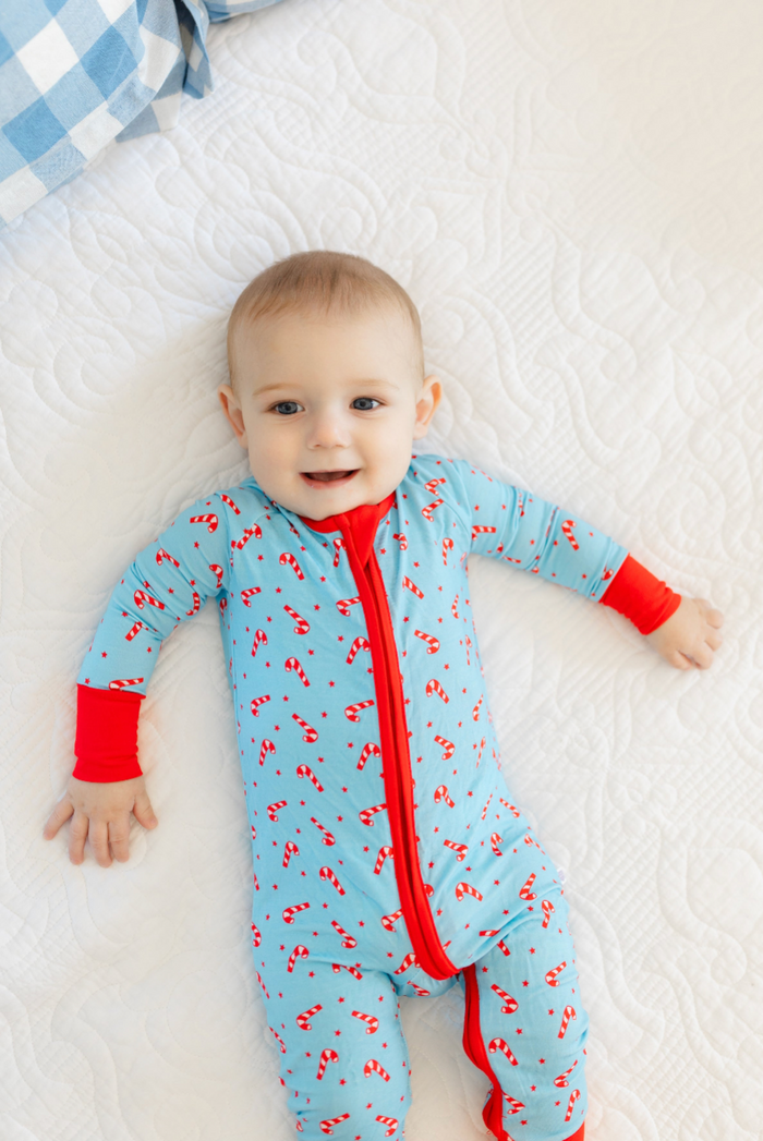 A baby lying on a white quilted mattress wearing a light blue footless sleeper with a red front zipper, red cuffs, and a red candy cane pattern.