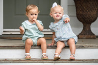 Two toddlers sitting on wooden porch steps wearing matching light blue gingham and floral dresses, with a large decorative urn visible in the background.