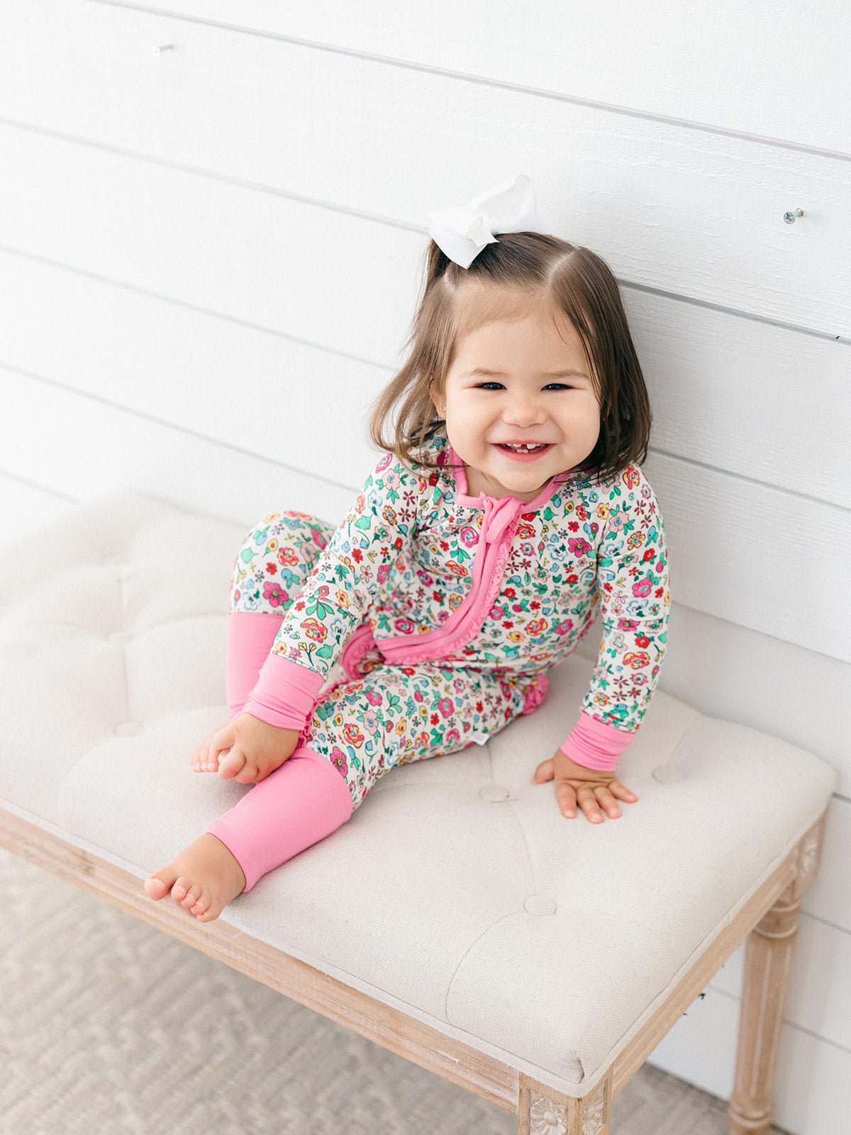 A smiling toddler girl sits on a tufted bench wearing a floral print zip-up sleeper with bright pink leg warmers and a white hair bow, set against a white wood-paneled wall.