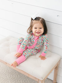 A smiling toddler girl sits on a tufted bench wearing a floral print zip-up sleeper with bright pink leg warmers and a white hair bow, set against a white wood-paneled wall.