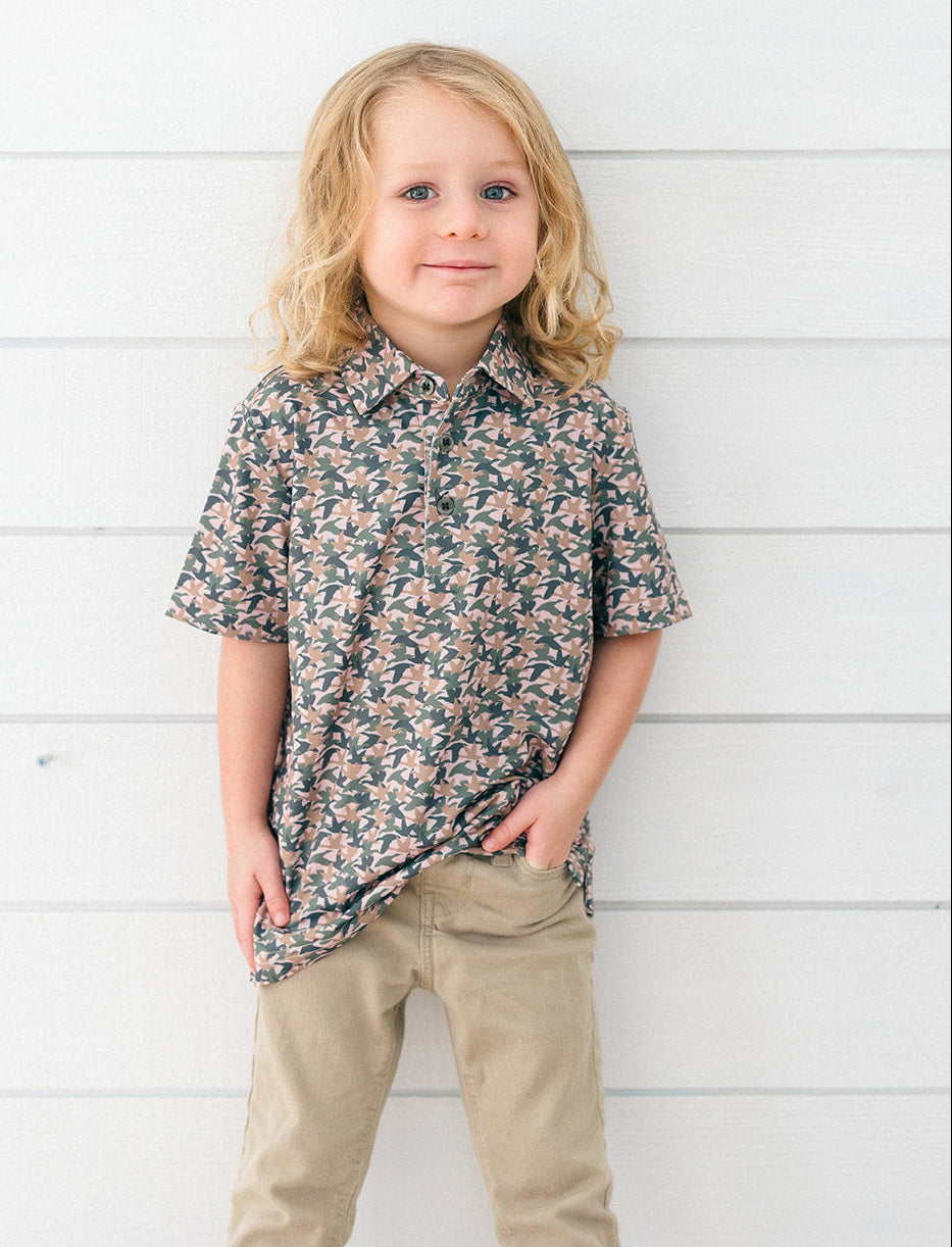 A young child with blonde hair stands against a white horizontal wood plank wall, wearing a short-sleeve button-up shirt with a dense floral pattern in dark green, pink, and beige, paired with khaki pants.