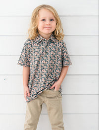 A young child with blonde hair stands against a white horizontal wood plank wall, wearing a short-sleeve button-up shirt with a dense floral pattern in dark green, pink, and beige, paired with khaki pants.