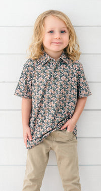A young child with blonde hair stands against a white horizontal wood plank wall, wearing a short-sleeve button-up shirt with a dense floral pattern in dark green, pink, and beige, paired with khaki pants.
