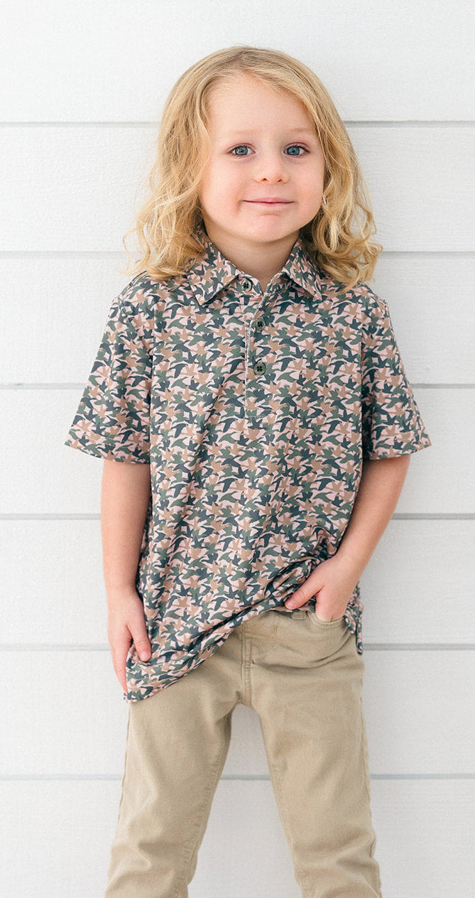 A young child with blonde hair stands against a white horizontal wood plank wall, wearing a short-sleeve button-up shirt with a dense floral pattern in dark green, pink, and beige, paired with khaki pants.