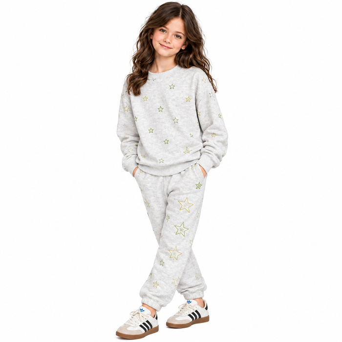 A young girl modeling a light gray heathered two-piece sweatshirt and jogger set, decorated with scattered gold star patterns. She is wearing white sneakers with black stripes, set against a plain white studio background.