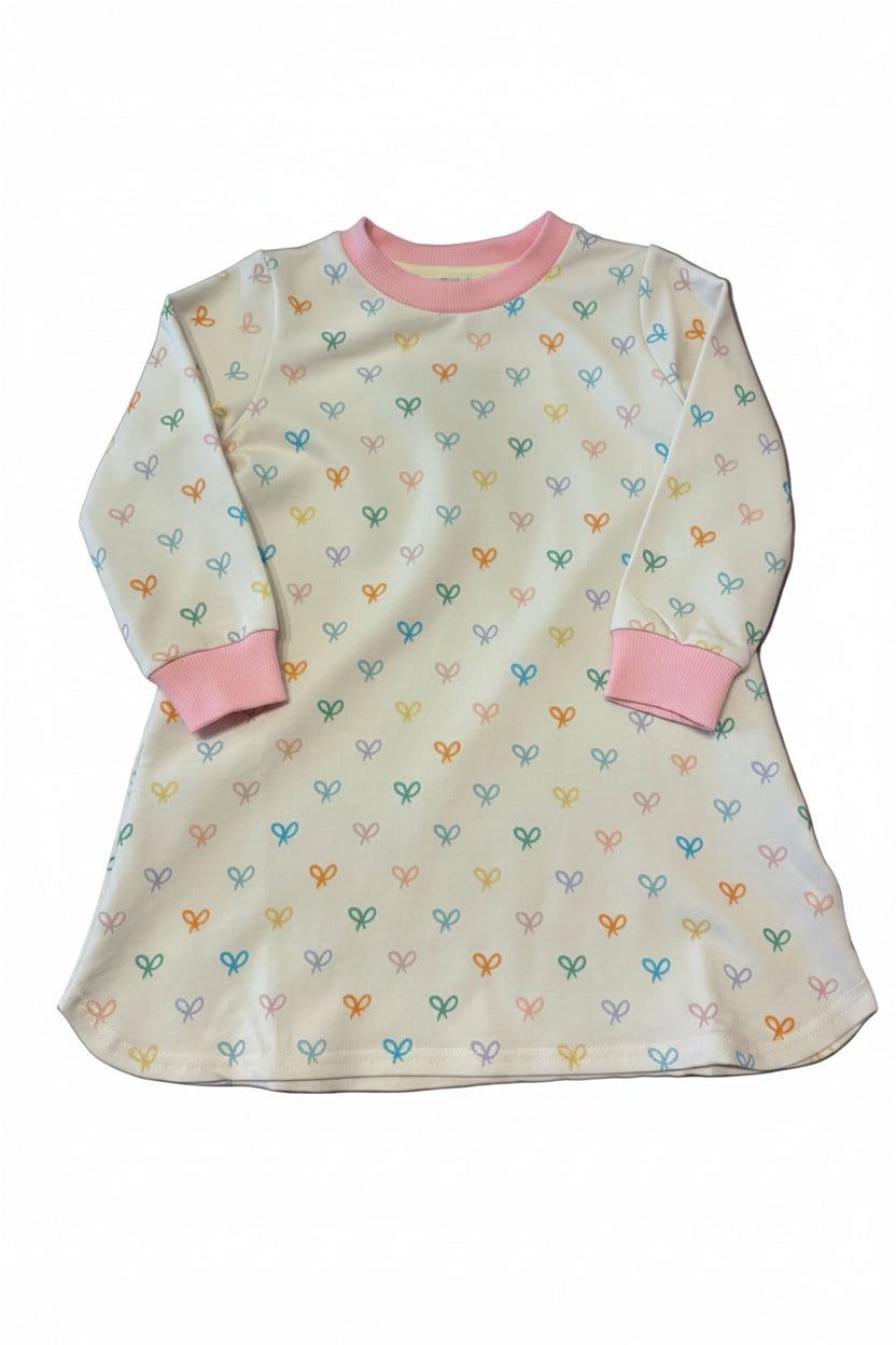 A long-sleeve baby tunic dress in cream with a colorful heart pattern and pink trim, laid flat on a gray surface.