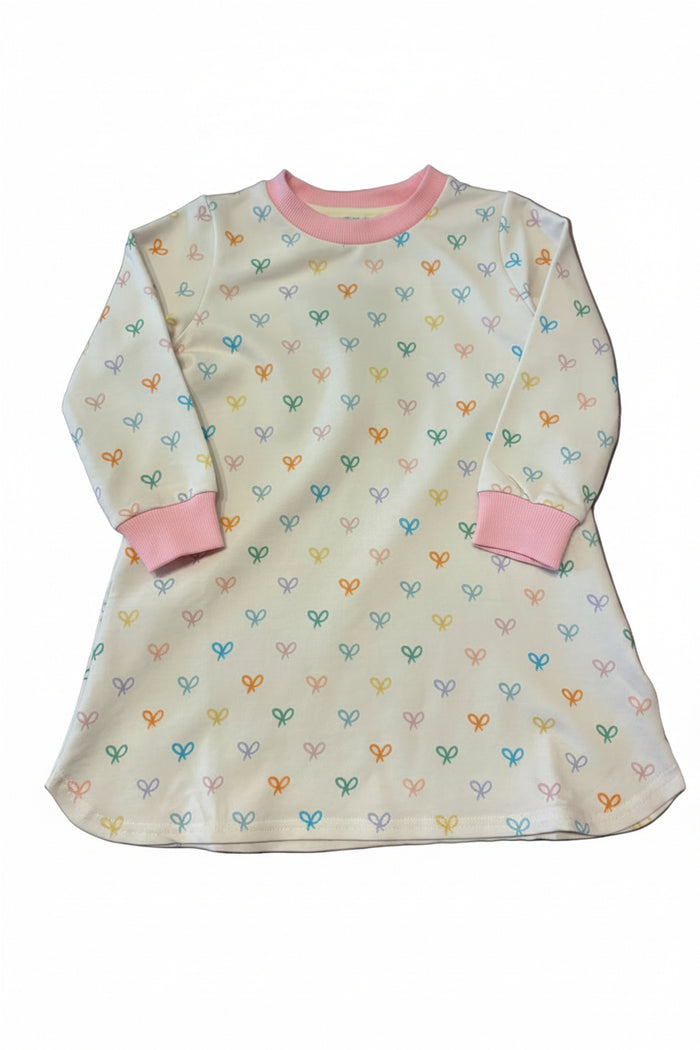 A long-sleeve baby tunic dress in cream with a colorful heart pattern and pink trim, laid flat on a gray surface.