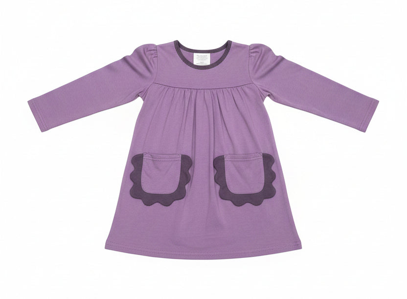 A purple long-sleeve baby dress with scalloped pocket edges, hanging on a clear plastic hanger in a retail store setting.