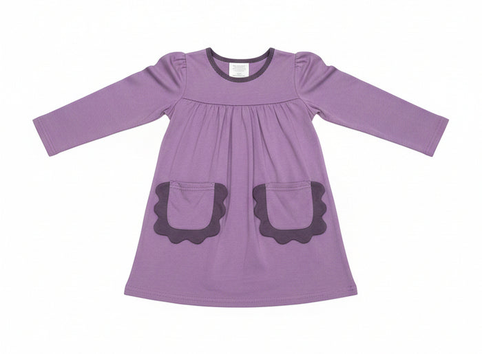 A purple long-sleeve baby dress with scalloped pocket edges, hanging on a clear plastic hanger in a retail store setting.