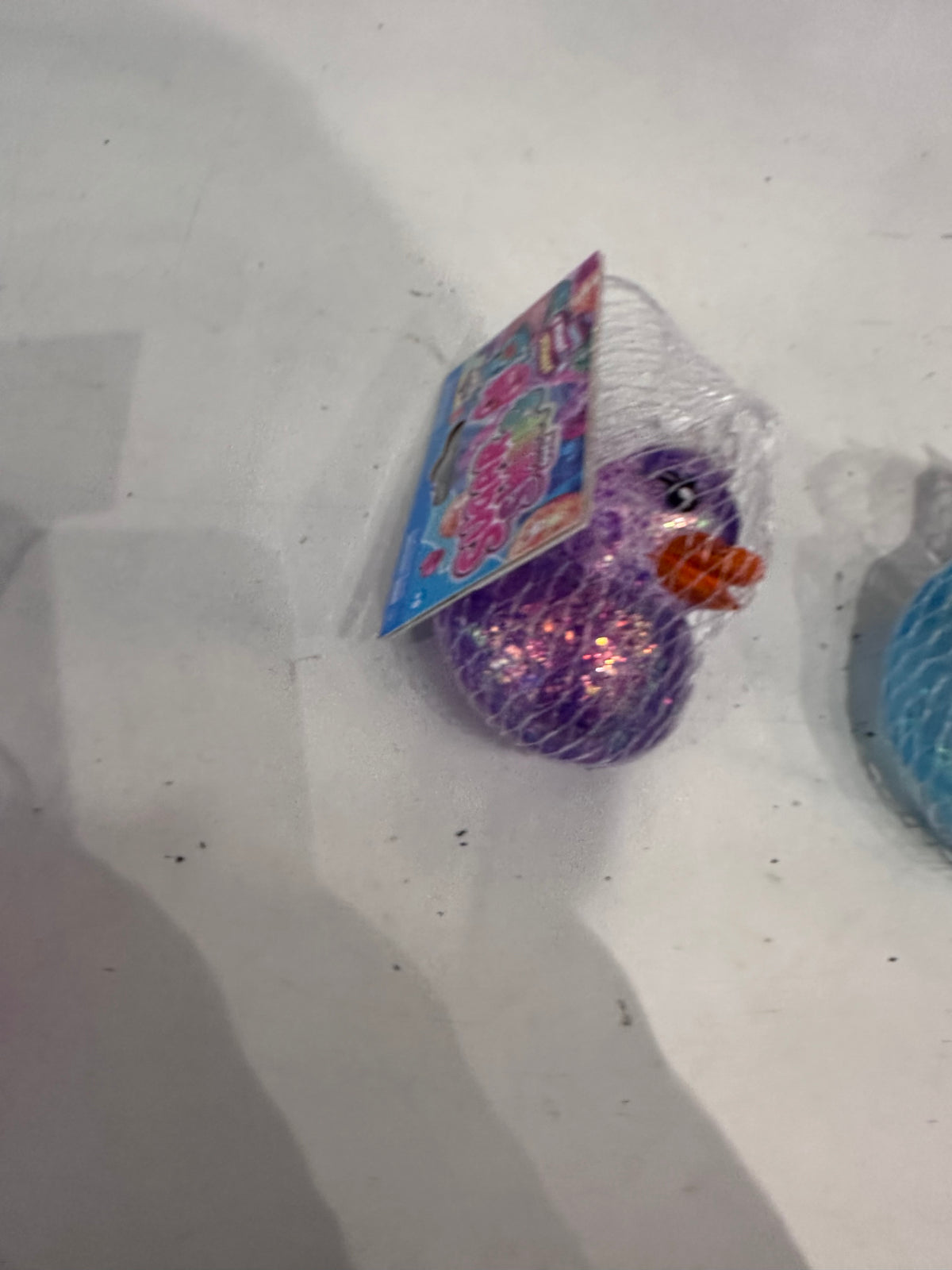 A small packaged toy duck in a clear mesh bag with a colorful printed tag, resting on a light surface. The duck is glittery purple with a bright orange beak.