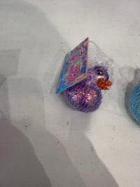A small packaged toy duck in a clear mesh bag with a colorful printed tag, resting on a light surface. The duck is glittery purple with a bright orange beak.