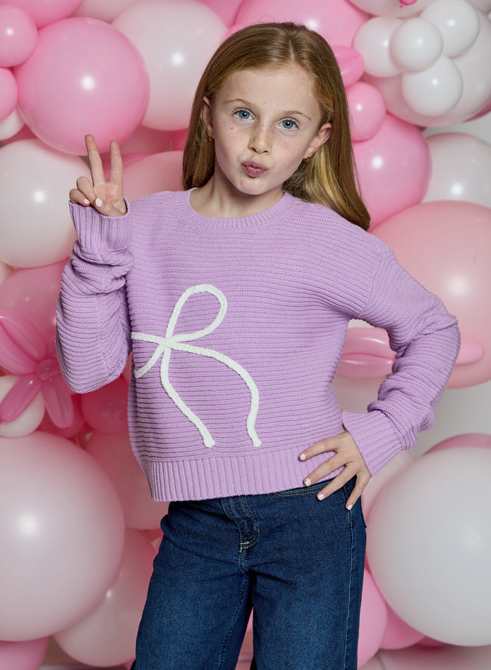 A young girl with light brown hair wearing a lavender ribbed knit sweater with a white bow graphic and blue jeans, posing in front of a pink and white balloon backdrop.