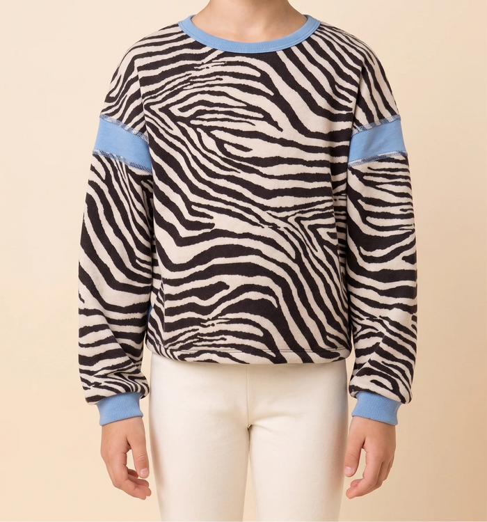 A young girl with a short brown bob haircut wearing a long-sleeve knit sweater with a black and beige zebra print, light blue ribbed collar, cuffs, and sleeve accents, paired with cream pants, against a solid beige background.