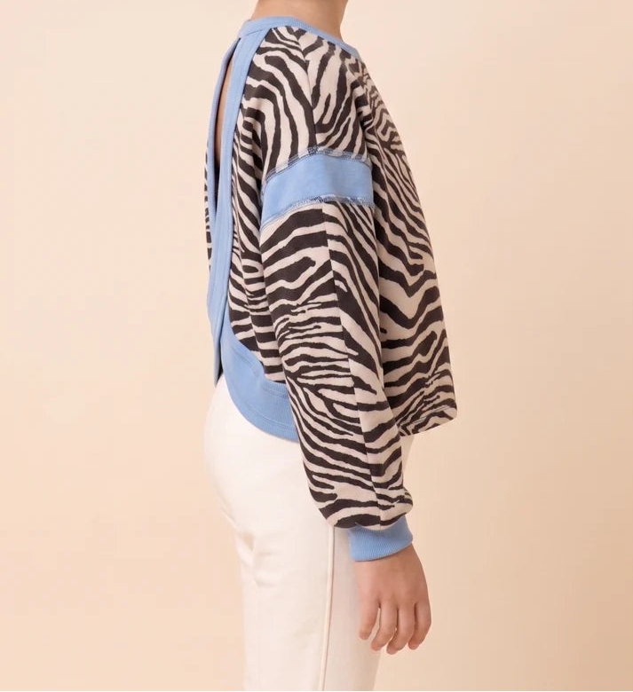 A child model shown in side profile wearing a long-sleeve knit top with a zebra print pattern, light blue color-blocked panels, and white pants against a solid light beige background.