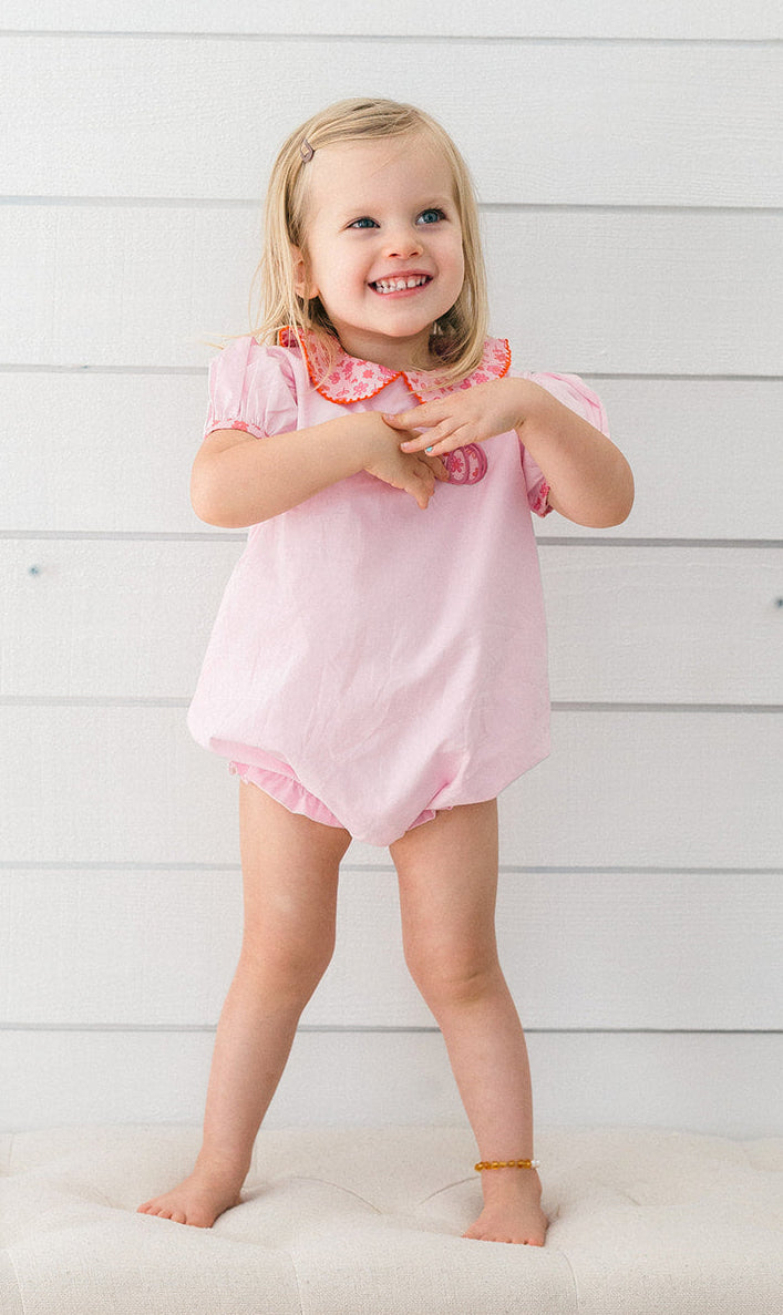 A young toddler girl with blonde hair stands smiling on a tufted cream bench against a white wood plank wall, wearing a light pink short-sleeve romper with a ruffled collar featuring a red and white pattern.