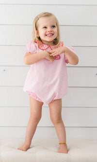 A young toddler girl with blonde hair stands smiling on a tufted cream bench against a white wood plank wall, wearing a light pink short-sleeve romper with a ruffled collar featuring a red and white pattern.