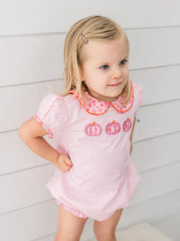 A young blonde girl wearing a light pink short-sleeve romper with three embroidered pink pumpkins and a floral collar, standing against a white paneled wall.