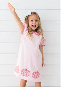 A young girl with blonde hair wearing a light pink dress with pink patterned pumpkin details at the hem and a white bow in her hair, standing against a white wood-paneled wall and waving.