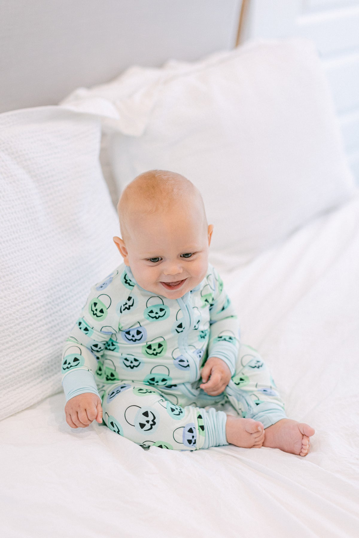 A baby sitting on a white bed wearing a light green and white long-sleeve footless pajama top with a green alien pattern.
