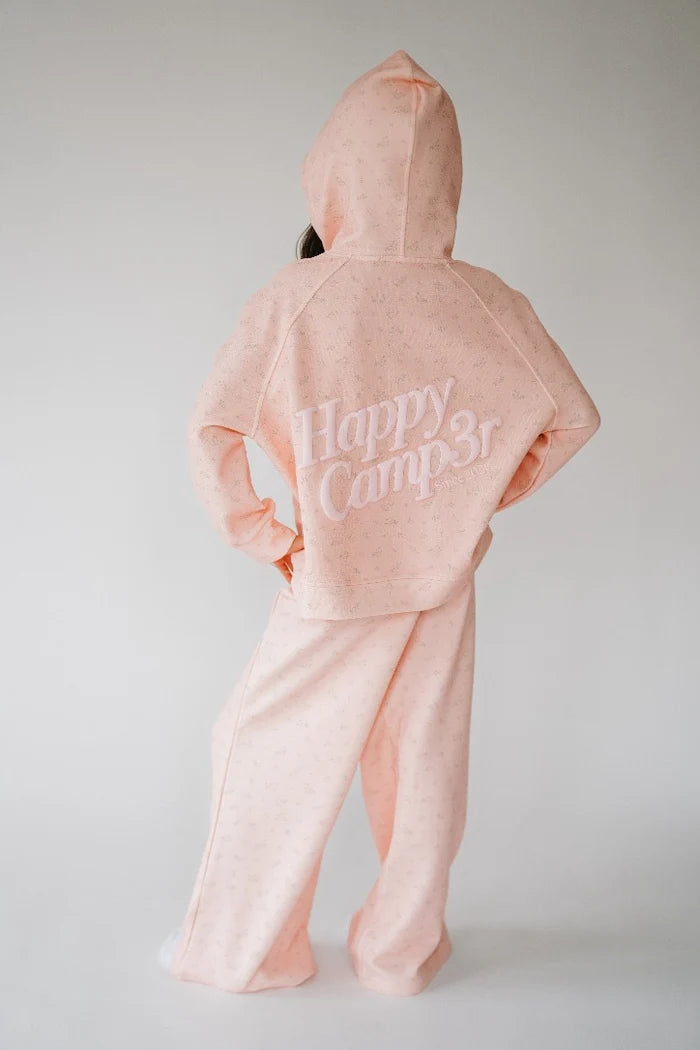 A child wearing a light pink hooded sweatshirt and matching sweatpants, shown from the back against a plain light gray studio background. The hoodie has 'Happy Camp3r' printed in white across the back.