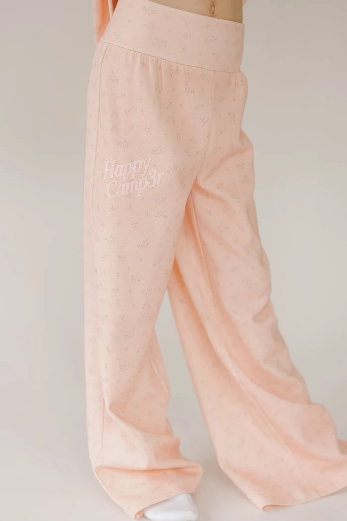 A person wearing light peach-pink wide-leg lounge pants with a subtle all-over floral print and 'Happy Camp3r' embroidered on the left thigh, shown against a plain light background.