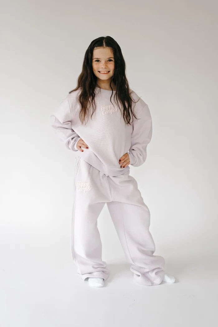 A young girl with long dark hair stands in a studio wearing a light lavender two-piece lounge set consisting of a sweatshirt and matching sweatpants. The set features subtle white embroidered text on the chest and thigh.