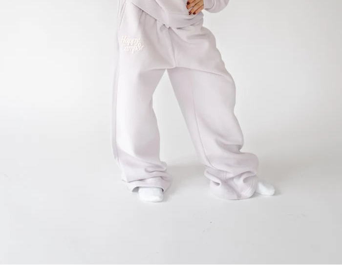 A young girl with long dark hair stands in a studio wearing a light lavender two-piece lounge set consisting of a sweatshirt and matching sweatpants. The set features subtle white embroidered text on the chest and thigh.