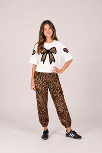 A young girl modeling a two-piece outfit consisting of a white t-shirt with a black bow and leopard print sleeve patches, paired with matching leopard print jogger pants, against a solid light gray background.