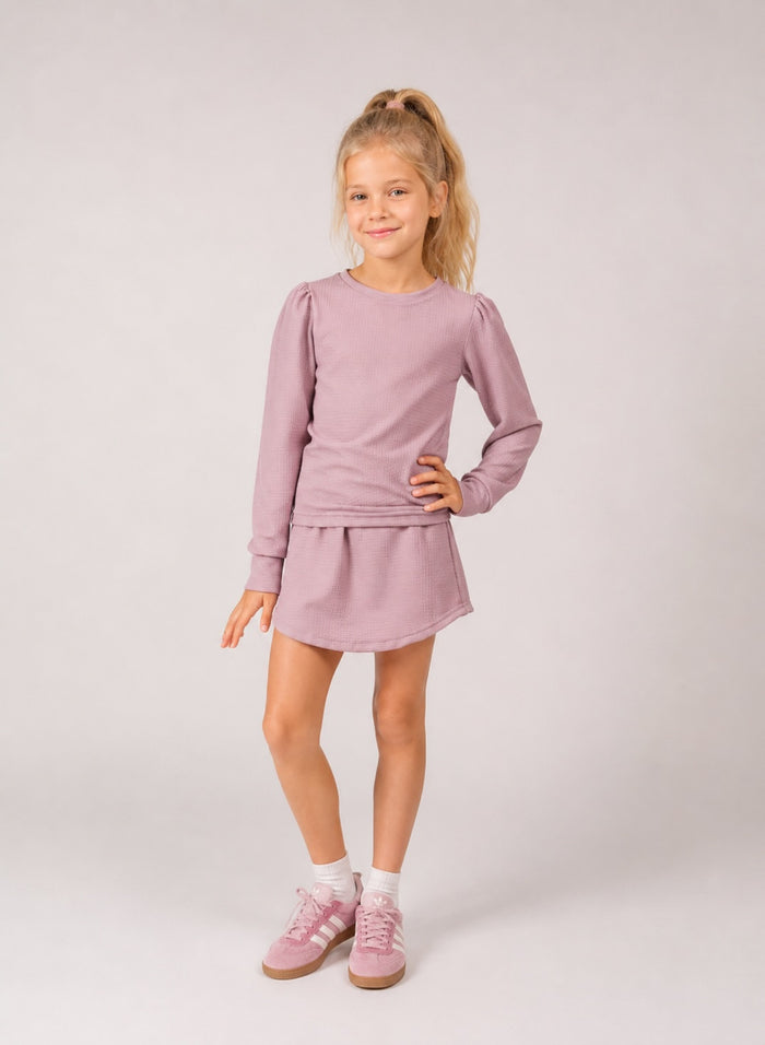 A young girl with blonde hair in a ponytail stands against a light gray studio background wearing a dusty pink long-sleeve top and matching mini skirt set, paired with white socks and pink sneakers.
