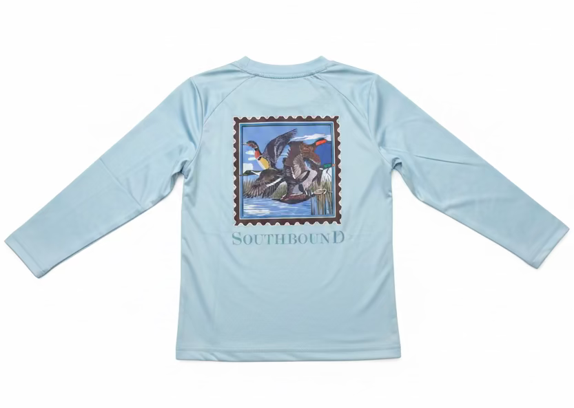 A light blue long-sleeve shirt with a large duck hunting graphic on the back, held up by a hand. The graphic features a stamp-like border with ducks in a marsh and the word 'SOUTHBOUND' printed below.