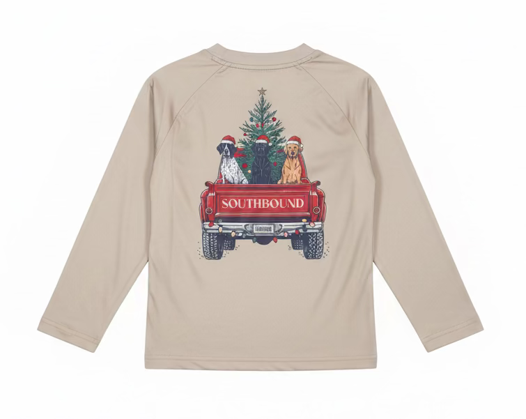 A light beige long-sleeve t-shirt held up on a hanger by a person's hand. The back of the shirt features a graphic print of three dogs wearing Santa hats sitting in a red pickup truck carrying a Christmas tree, with the word 'SOUTHBOUND' printed on the truck bed.