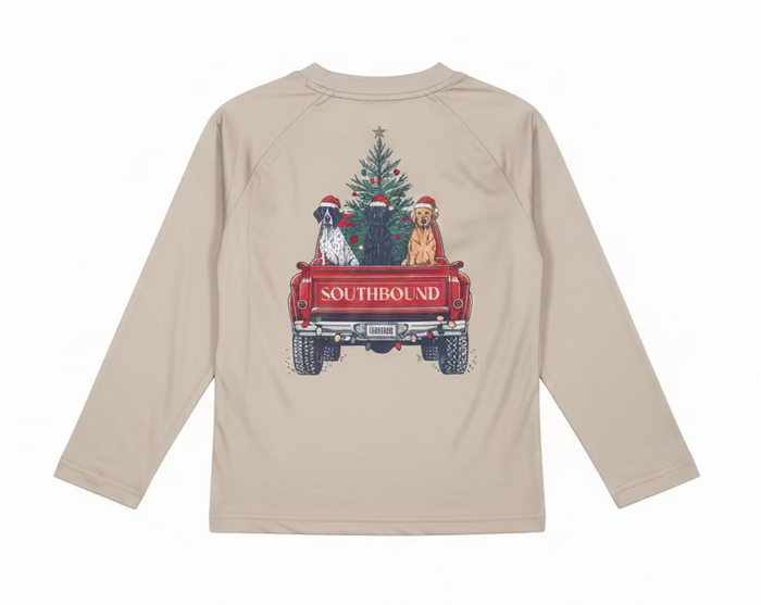 A light beige long-sleeve t-shirt held up on a hanger by a person's hand. The back of the shirt features a graphic print of three dogs wearing Santa hats sitting in a red pickup truck carrying a Christmas tree, with the word 'SOUTHBOUND' printed on the truck bed.