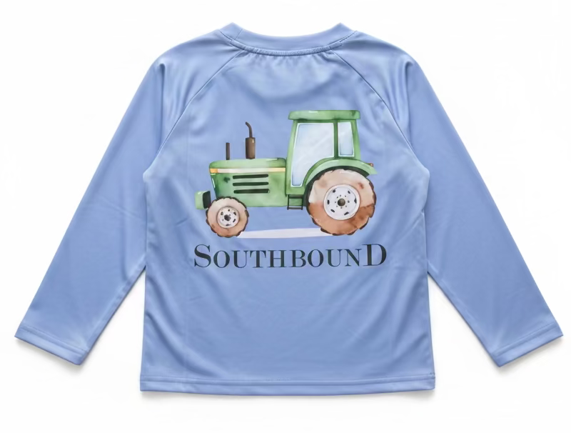 A light blue long-sleeve raglan shirt on a hanger, held by a hand with gold bracelets. The shirt features a green tractor graphic and the text 'SOUTHBOUND' printed on the back.
