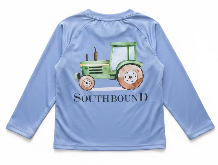 A light blue long-sleeve raglan shirt on a hanger, held by a hand with gold bracelets. The shirt features a green tractor graphic and the text 'SOUTHBOUND' printed on the back.