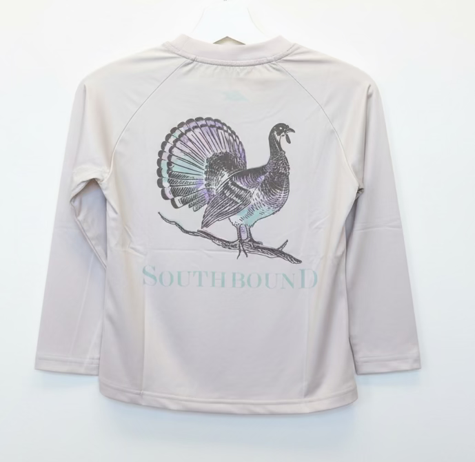 A child's long-sleeve raglan shirt in a light beige or off-white color, held up on a hanger by a person's hand. The back of the shirt features a large graphic of a turkey with iridescent glittery feathers and the word 'SOUTHBOUND' printed below in a matching glittery font.