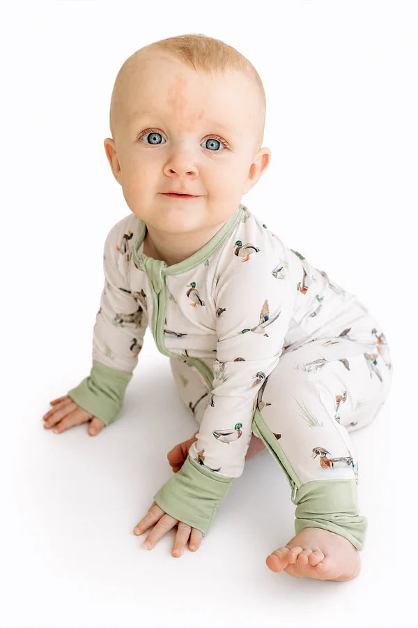 A baby wearing a long-sleeve footless pajama romper with a duck print and sage green cuffs, crawling on a white background.