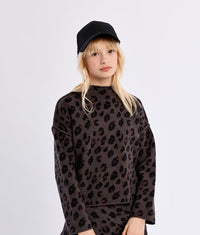 A young girl with blonde hair wearing a black baseball cap, a dark brown and black leopard-print long-sleeve top, and matching shorts, standing against a light gray studio background.