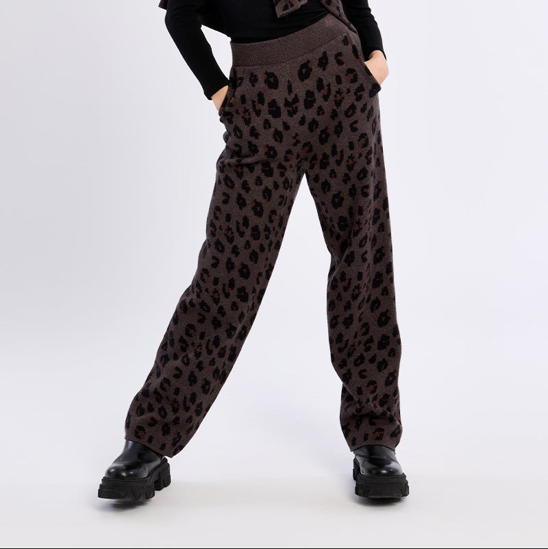 A model posing in a studio setting wearing a black long-sleeve top with a ruffled collar, brown leopard-print pants, and black shoes.