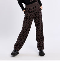 A model posing in a studio setting wearing a black long-sleeve top with a ruffled collar, brown leopard-print pants, and black shoes.